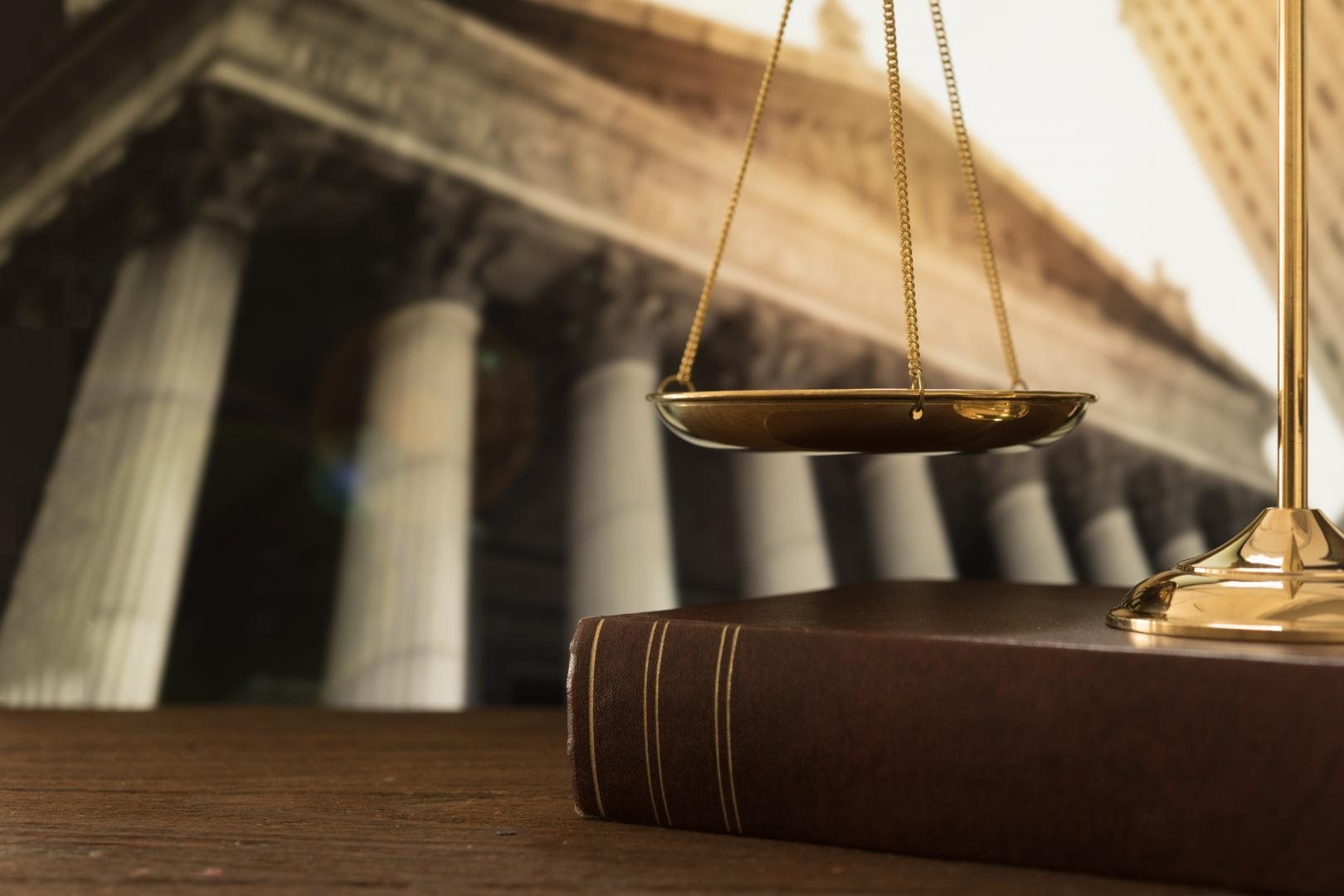 Un plateau de balance de la justice en laiton doré est suspendu au-dessus d’un livre relié de cuir brun, devant un bâtiment à colonnes flou.