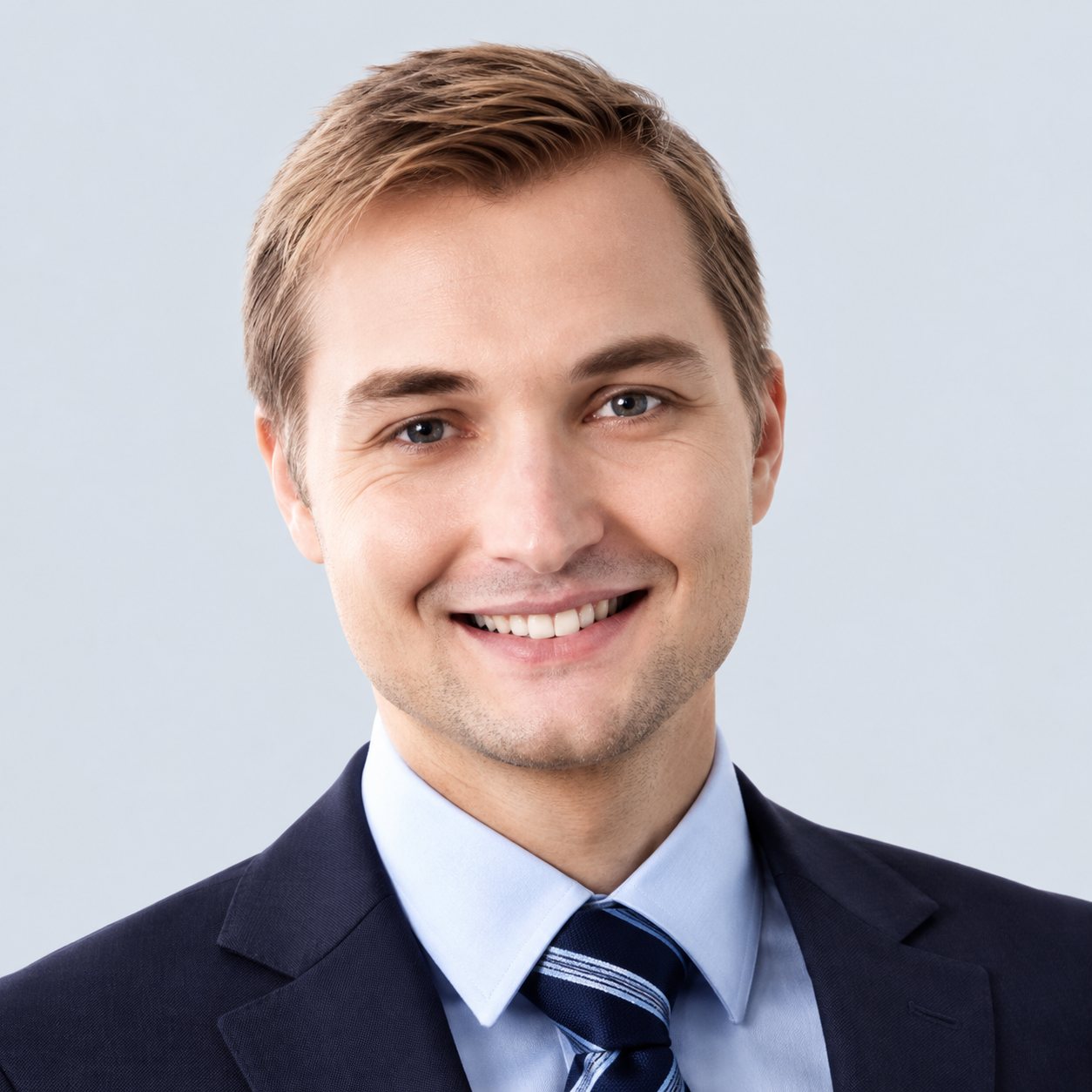 Portrait d’un jeune homme souriant, vêtu d’un costume sombre, d’une chemise blanche et d’une cravate rayée, devant un fond clair.
